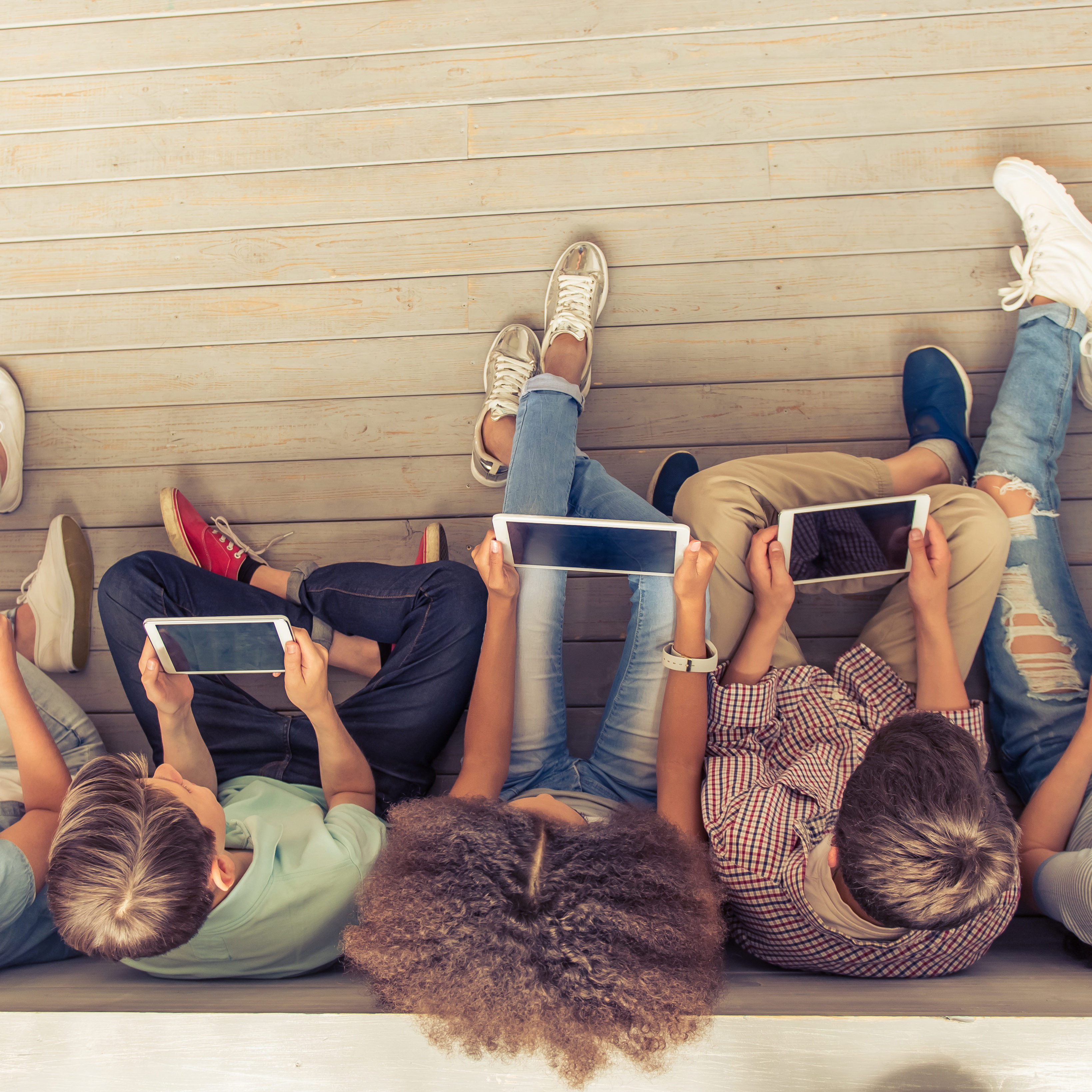 Top view of group of teenage boys and girls using tablets while sitting in row on wooden floor